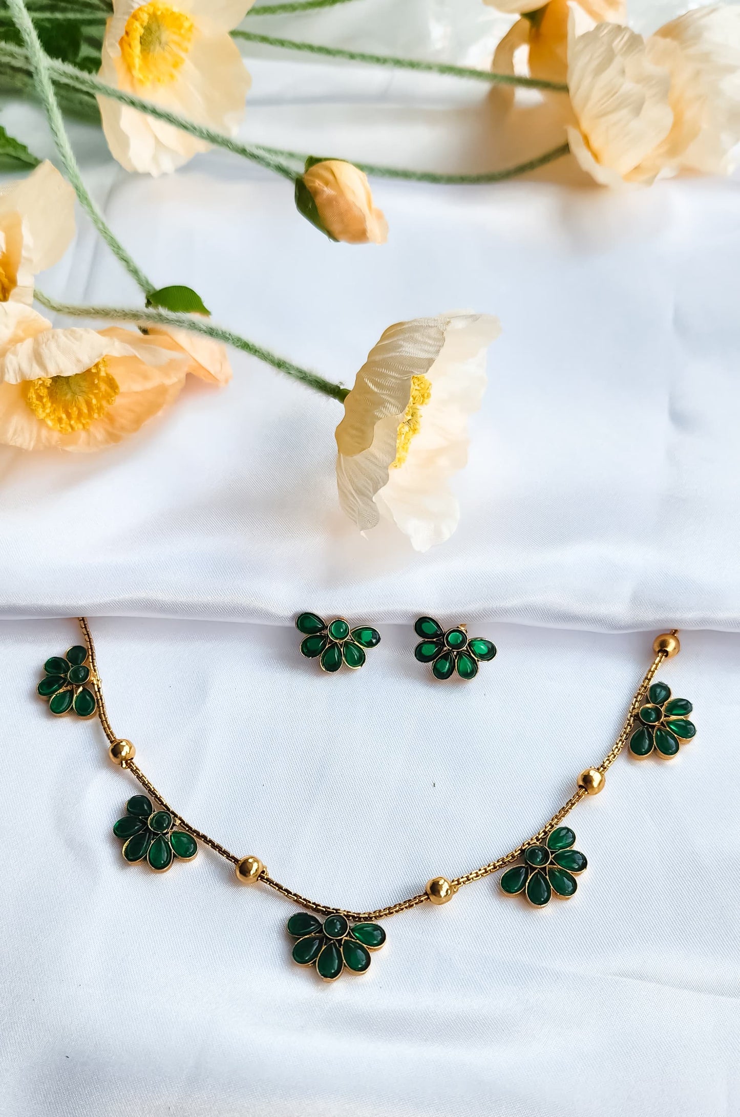 Green floral necklace with matching stud earrings displayed on white fabric with soft flowers in the background.