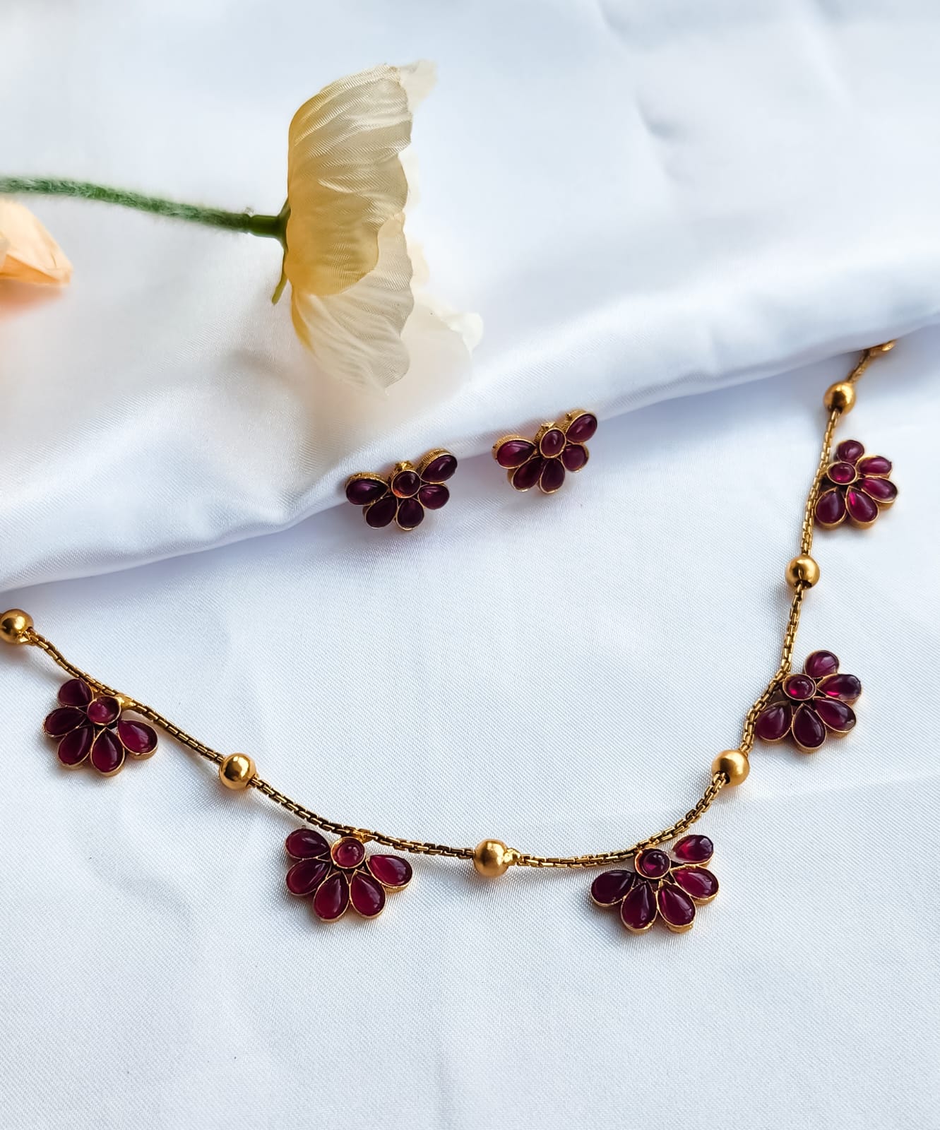 Pink floral necklace with matching stud earrings styled on white fabric, complemented by peach flowers in the background.