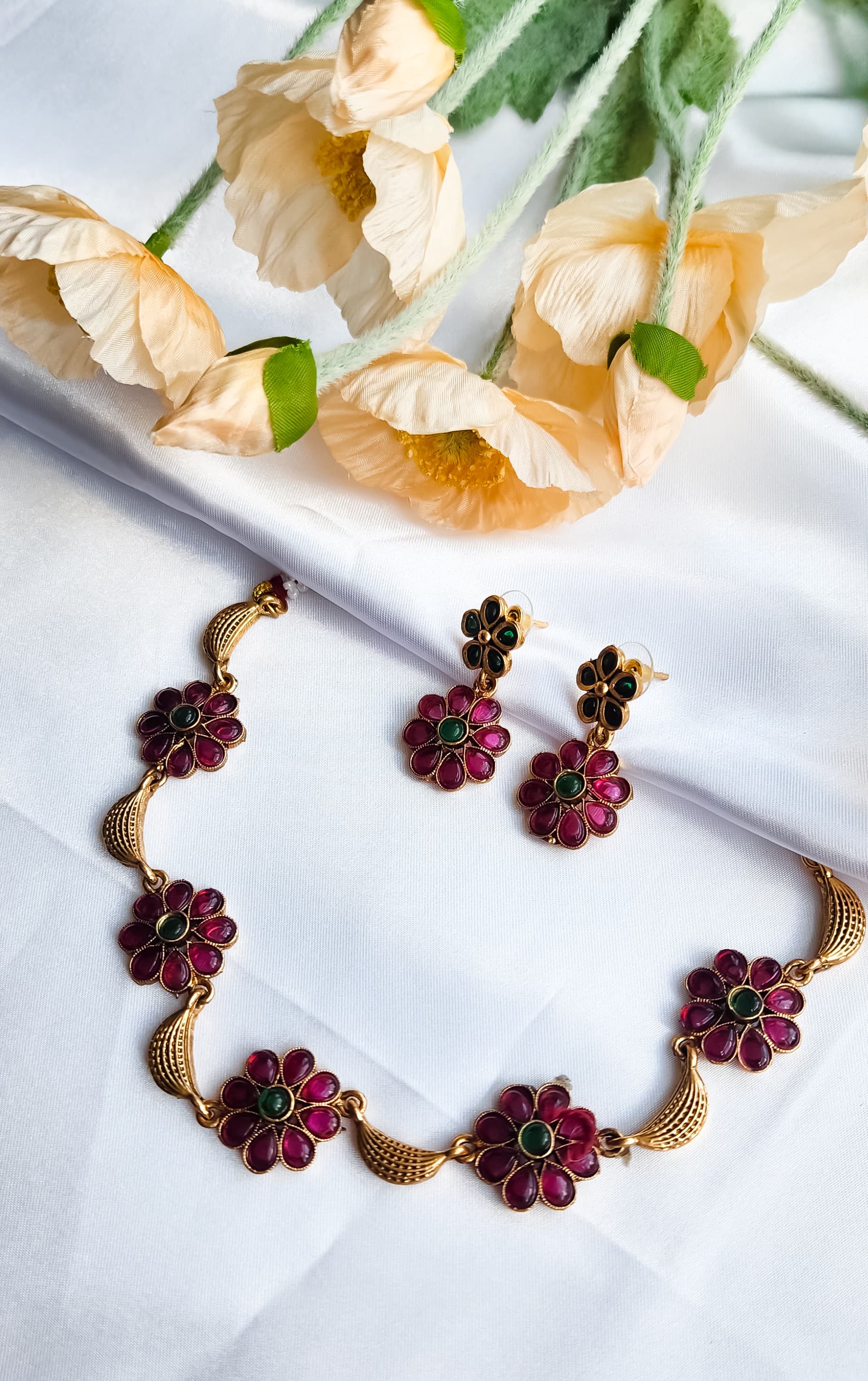 Ruby-red and green floral kundan necklace set with gold detailing displayed elegantly on fabric.