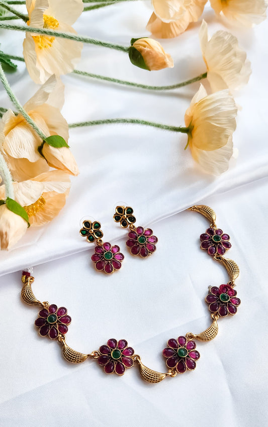 A floral ruby and green kundan necklace with matching earrings placed on a white background with peach flowers.