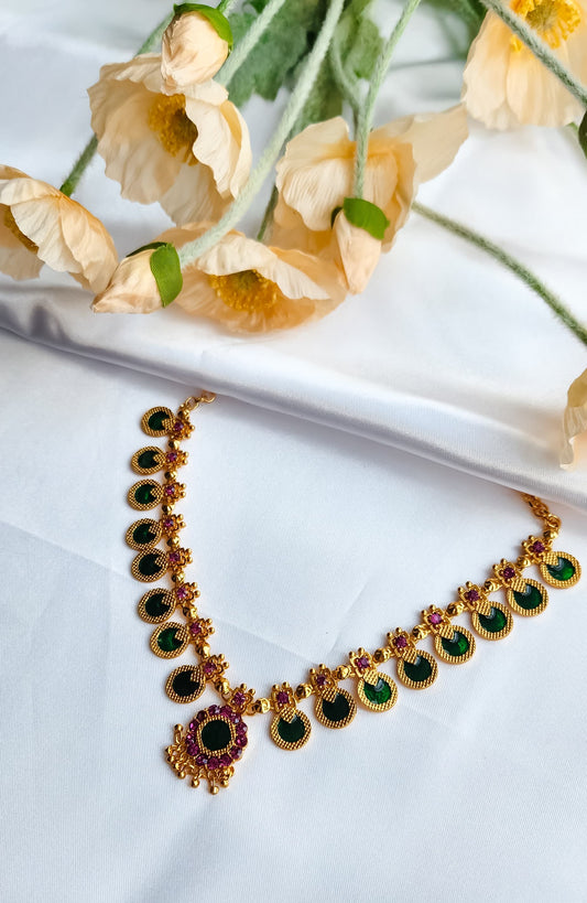 Traditional Lakshmi Kasu style choker necklace with green enamel kasu motifs and ruby floral accents, placed on white fabric with floral props.