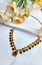 Traditional Lakshmi Kasu style choker necklace with green enamel kasu motifs and ruby floral accents, placed on white fabric with floral props.