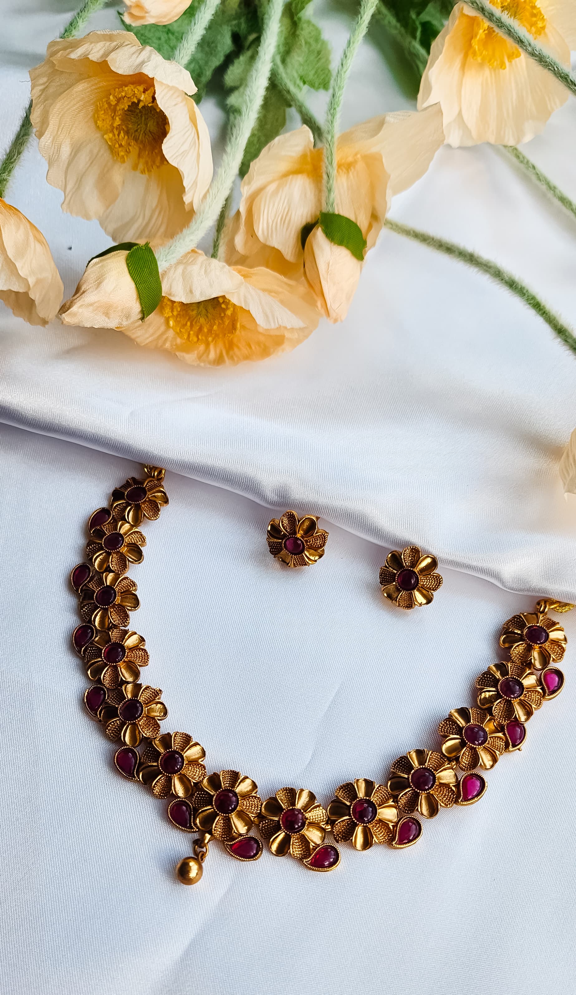 Close-up of a ruby red floral necklace and earrings set arranged on a white background, featuring intricate gold-toned flowers with red stones.
