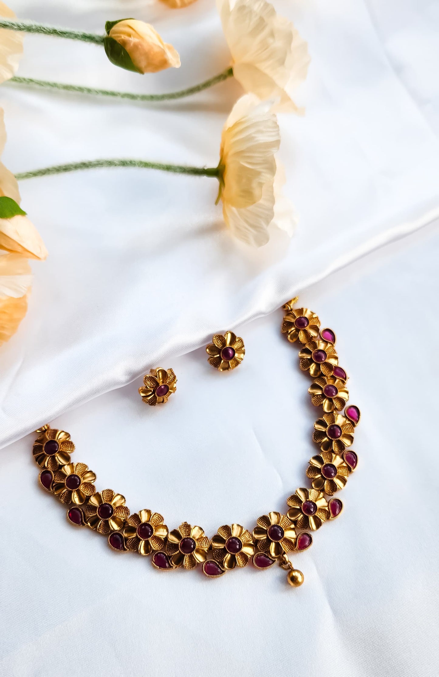 Traditional floral motif necklace with ruby red stones and golden drop pendant, paired with matching stud earrings, styled with decorative flowers.
