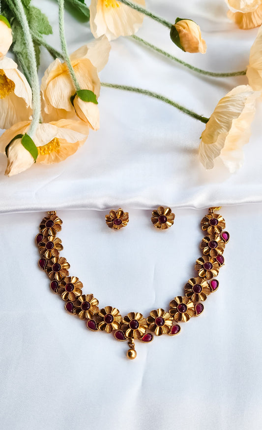 Golden floral motif necklace with ruby red stones and matching floral stud earrings, displayed on white fabric with cream flowers in the background.