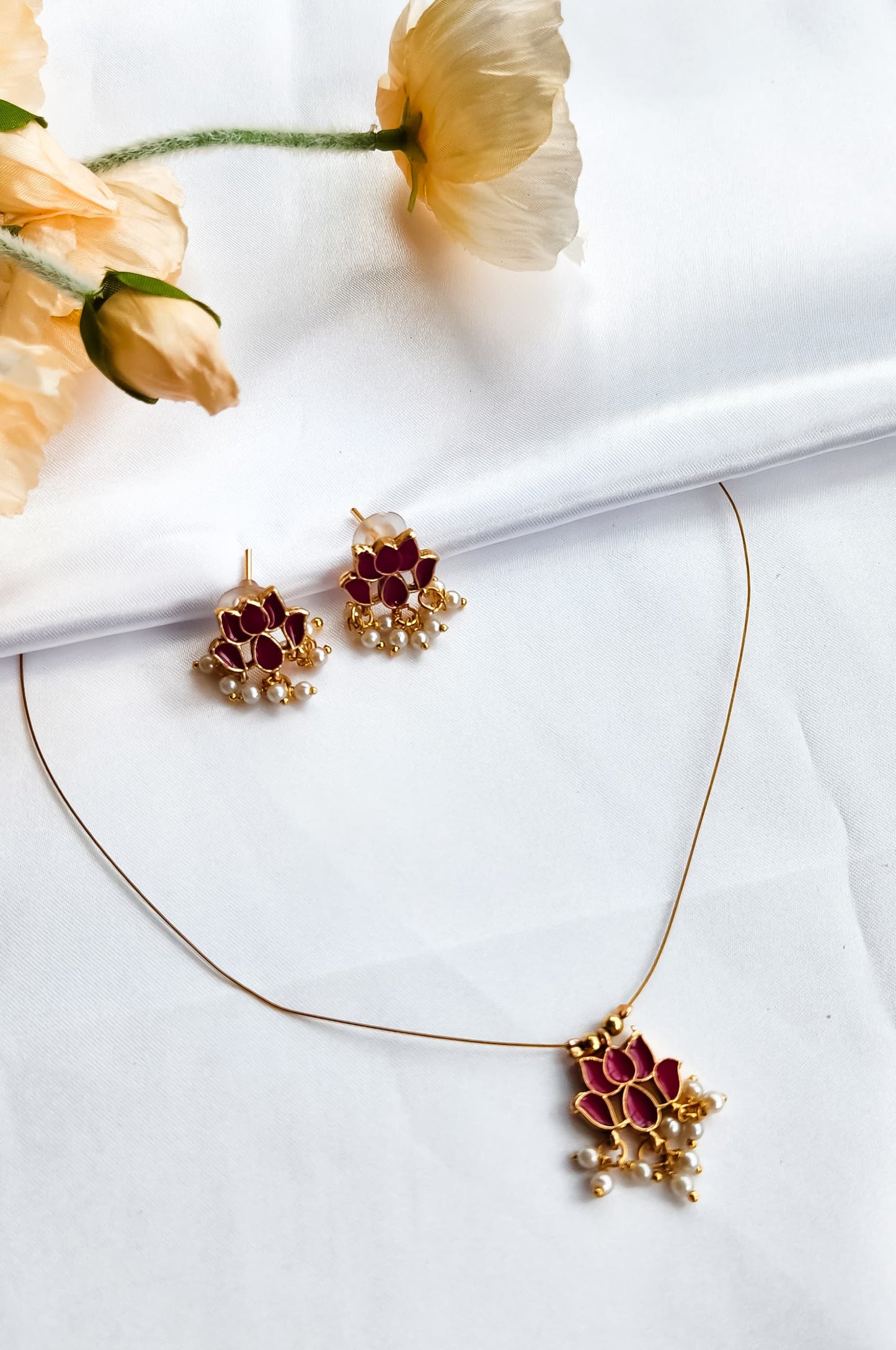 Red lotus design necklace with matching earrings adorned with pearl drops, placed on a white fabric background with flowers.