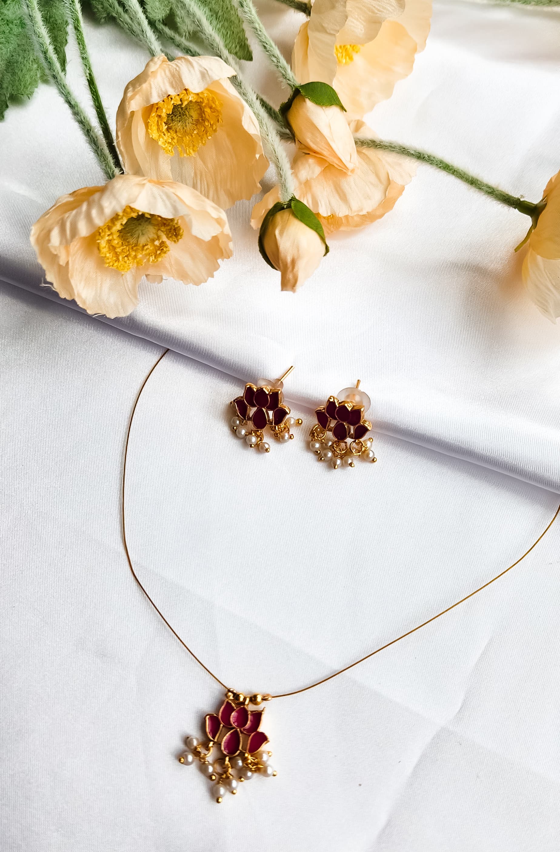 Red lotus design necklace with matching earrings adorned with pearl drops, placed on a white fabric background with flowers.