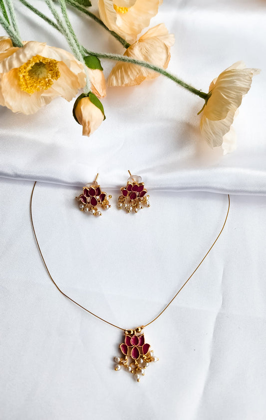 Red lotus design necklace with matching earrings adorned with pearl drops, placed on a white fabric background with flowers.