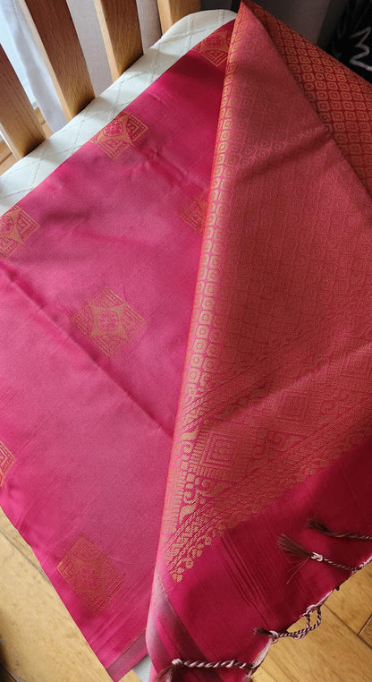 Deep pink soft silk saree with gold zari motifs and geometric woven pallu, displayed folded with tassels for festive and traditional wear.