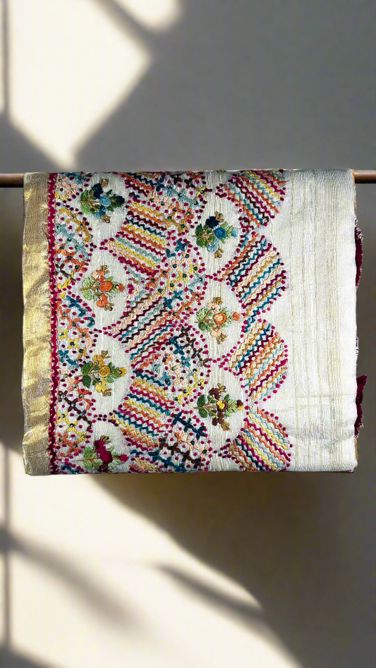 Off-white semi tussar silk saree with multicolor Resham thread embroidery, golden zari border, and maroon tassels.
