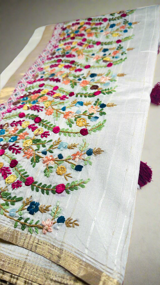 Off-white semi tussar silk saree with multicolor Resham thread embroidery, golden zari border, and maroon tassels.
