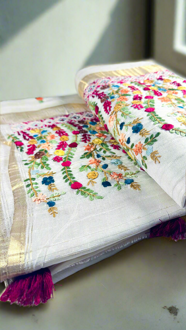 Off-white semi tussar silk saree with multicolor Resham thread embroidery, golden zari border, and maroon tassels.
