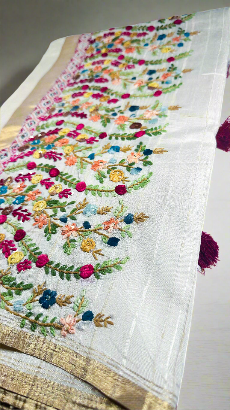 Off-white semi tussar silk saree with multicolor Resham thread embroidery, golden zari border, and maroon tassels.
