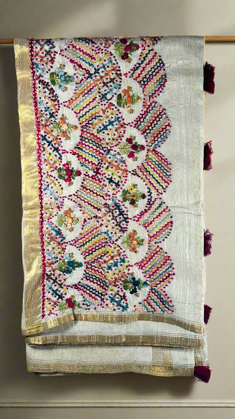 Off-white semi tussar silk saree with multicolor Resham thread embroidery, golden zari border, and maroon tassels.
