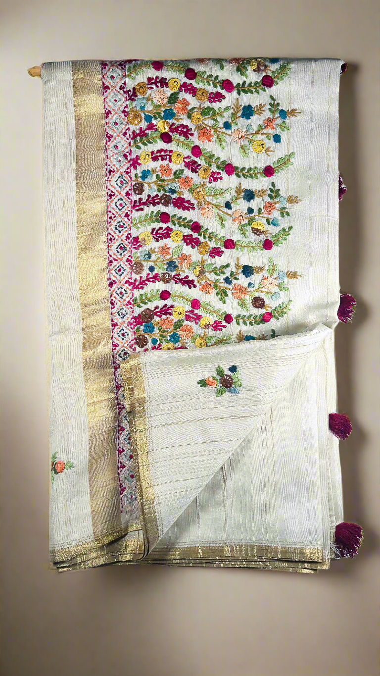 Off-white semi tussar silk saree with multicolor Resham thread embroidery, golden zari border, and maroon tassels.
