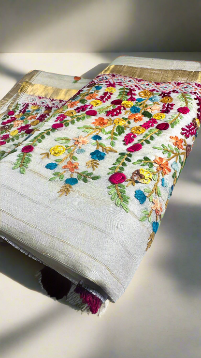 Off-white semi tussar silk saree with multicolor Resham thread embroidery, golden zari border, and maroon tassels.
