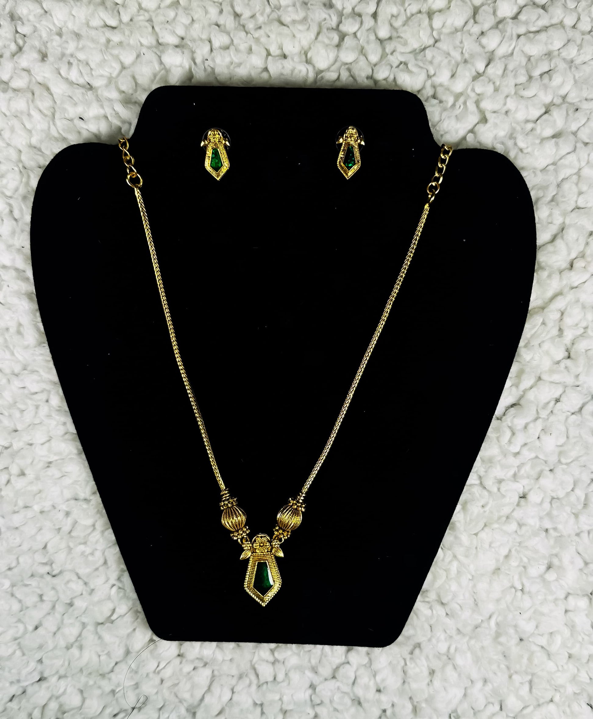 Gold plated geometric necklace and earrings jewelry set with emerald green diamond-shaped pendants displayed on black velvet jewelry stand
