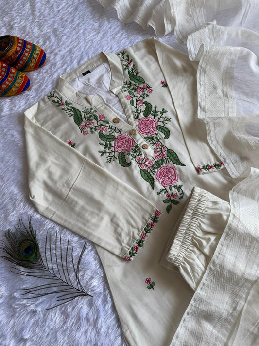 Woman wearing a white khadi cotton salwar suit with pink floral embroidery, matching straight pants, and a fringed dupatta, standing indoors on a patterned rug.