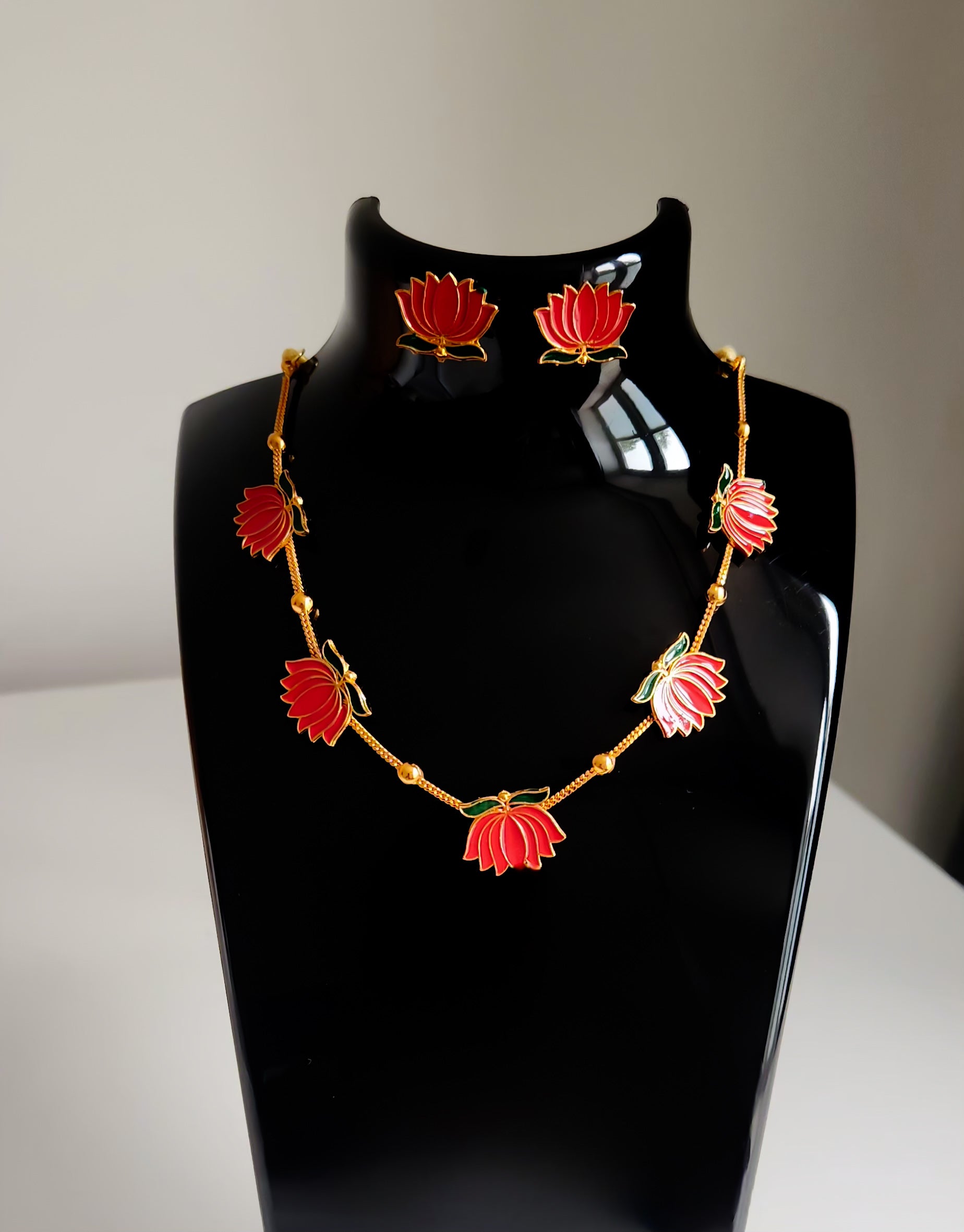 Red lotus enamel necklace with golden beads and matching earrings on black display.
