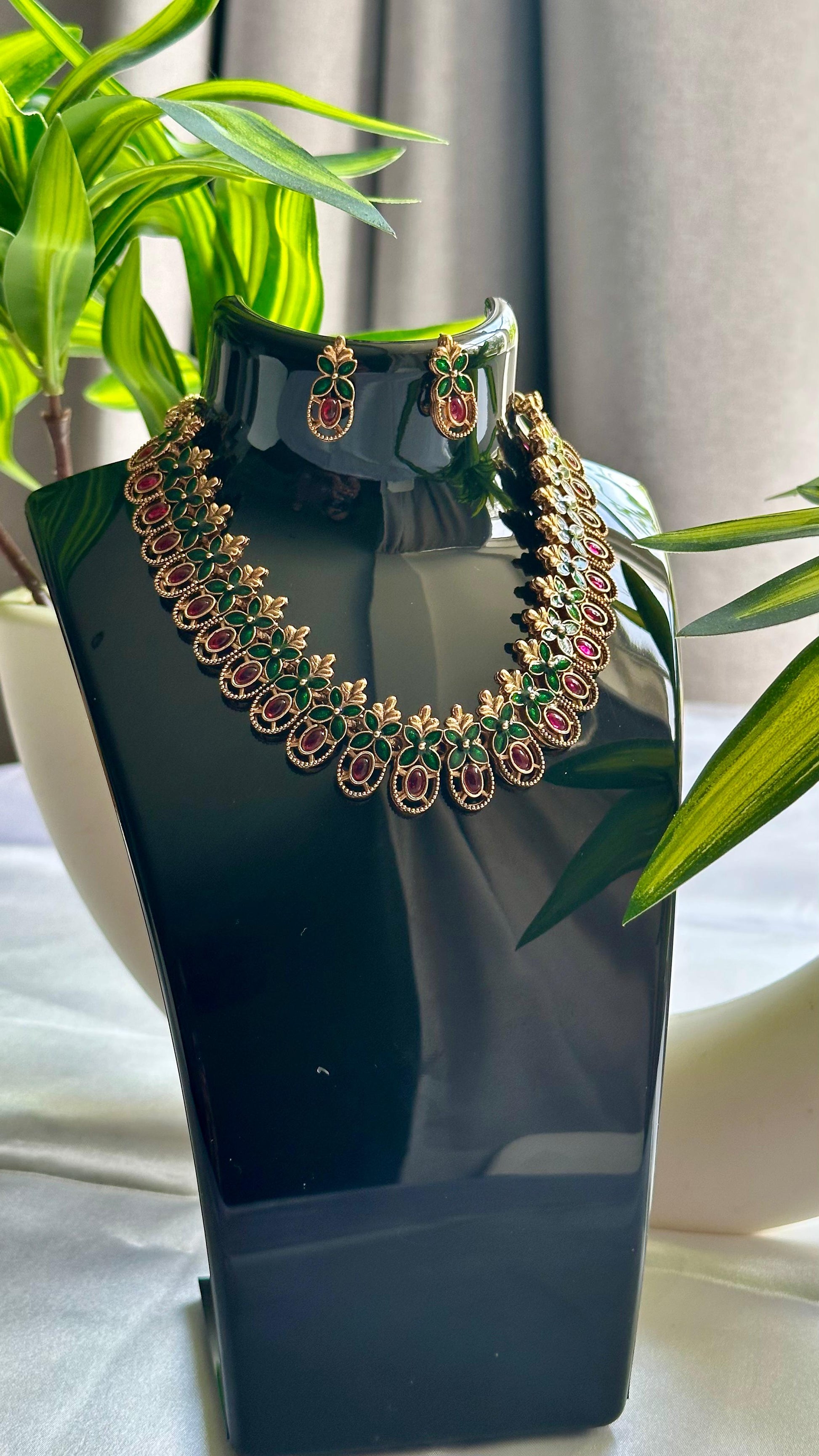 Gold-plated necklace and earrings set featuring red teardrop stones with green enamel accents and intricate gold detailing, displayed on a black stand with green plants and a white vase in the background.
