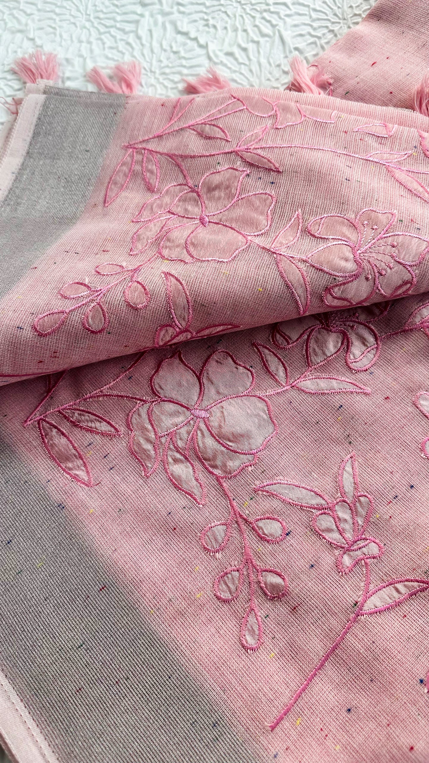 Soft pink linen saree with floral appliqué embroidery, speckled pattern, grey border, and pink tassels.
