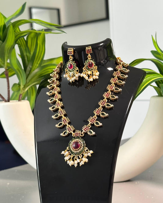 Traditional gold-plated necklace and earrings set featuring a central red stone pendant with pearl-like bead drops, displayed on a glossy black bust with green plants and white pots in the background.
