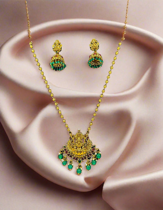 Traditional Indian temple jewelry set with gold Lakshmi pendant necklace and emerald green jhumka earrings displayed on black velvet stand
