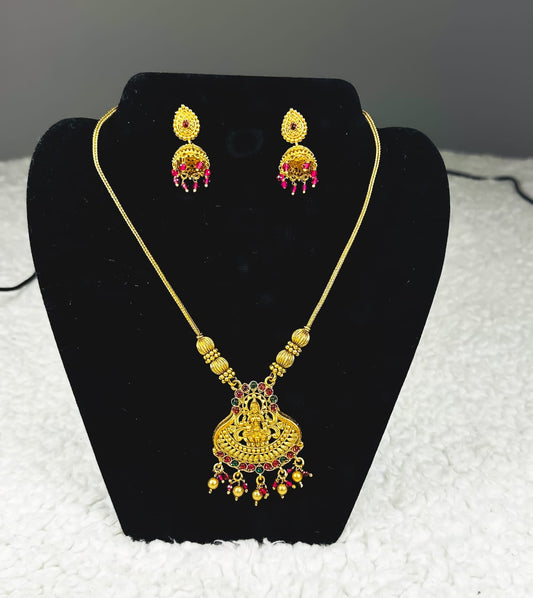 Traditional Indian temple jewelry set with ornate gold Lakshmi pendant necklace and ruby jhumka earrings featuring goddess motifs and temple craftsmanship displayed on black velvet stand